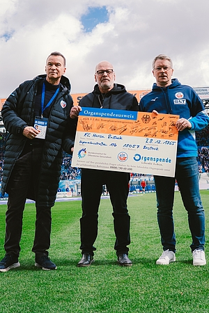 Drei Männer stehen auf dem Rasen des Ostseestadions und präsentieren gemeinsam einen überdimensionalen, symbolischen Organspendeausweis des F.C. Hansa Rostock. Der Ausweis ist auf den Namen des Vereins ausgestellt und trägt zahlreiche Unterschriften.